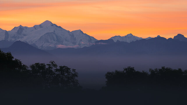 View of sunrise over the snow-capped Mont Blanc massif with silhouettes of trees in the foreground and a vibrant orange sky in Chamonix, Auvergne-Rhone-Alpes, France.