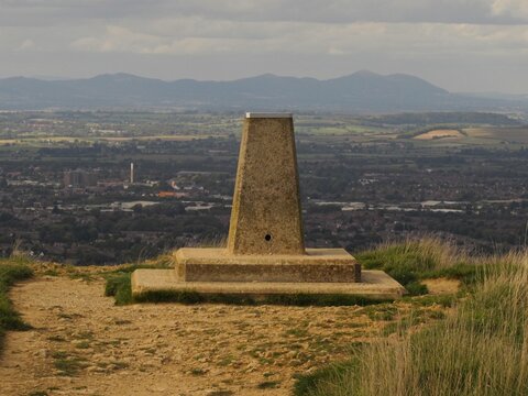 A concrete triangulation pillar stands on Painswick Beacon, UK overlooking Gloucester in the valley below, with the rolling Malvern Hills and mountains in the distance under a cloudy sky