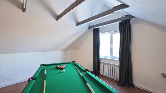Attic billiard room with green table, cues, balls, slanted ceiling, exposed beams, window with dark curtains, radiator, minimalist finish.
