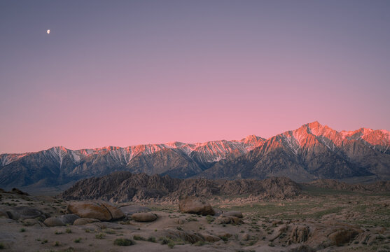 View of Alabama Hills with the snow-capped Sierra Nevada mountains under a pink sunset sky and a visible moon in Lone Pine, California, United States.