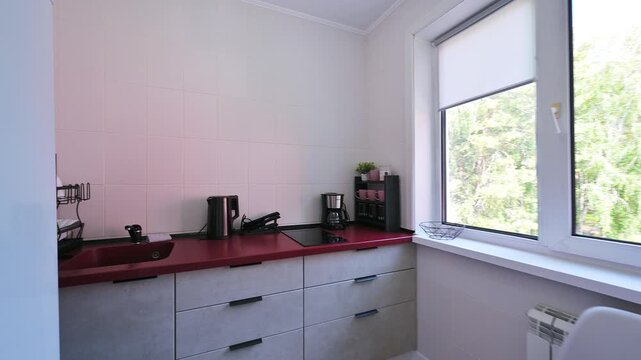 Compact kitchen nook with white cabinets, red countertop, black handles, window with roller blind, minimal clutter, functional layout.
