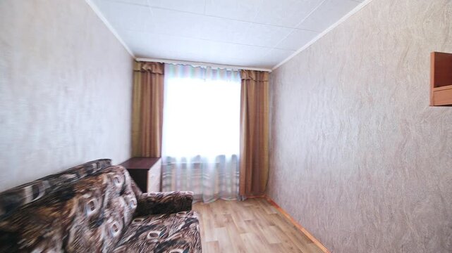 Compact room with patterned sofa, beige textured walls, wooden floor, window with layered curtains, simple built-in shelf, neutral interior.
