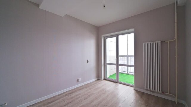 Spacious empty room with light wooden flooring, pale beige walls, white radiator, sliding glass door to balcony with green flooring.
