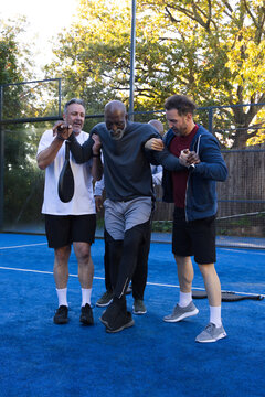 Male friends supporting senior player limping on blue court in sportswear with paddles and net