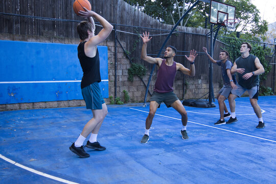 Diverse males in shorts playing basketball on blue court with hoop, shooter holding ball