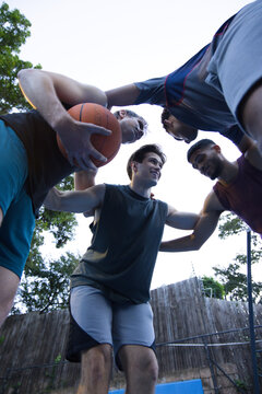 Four adult males huddling on outdoor court holding orange basketball by hoop, wearing sportswear