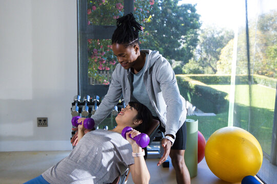 African American man spotting Asian woman pressing purple dumbbells on decline bench in home gym