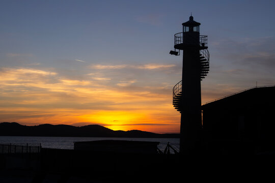 Coastal lighthouse silhouetted against a glowing sunset over calm water and distant hills