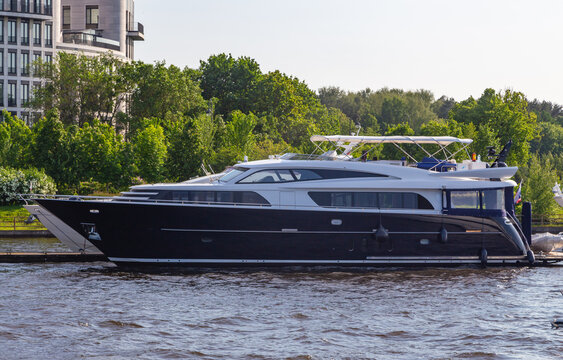Luxury yacht glides gently through calm water. Tree-lined riverbanks frame modern architecture. Smooth hull reflects sky and greenery. Serene moment suggests quiet luxury. Ideal for travel, leisure
