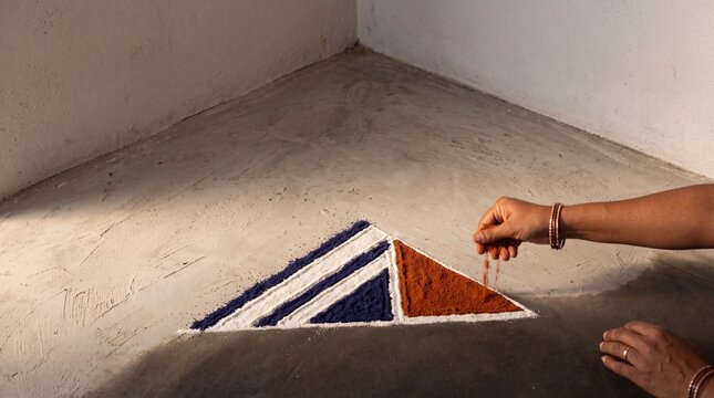 Close-up of a woman's hands wearing bangles creating a geometric triangular rangoli pattern with orange, blue, and white powder on a concrete floor