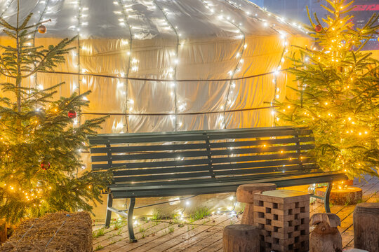 View of a festive Christmas market scene featuring a white yurt adorned with fairy lights, a dark wooden bench, and decorated Christmas trees on a wooden deck in Switzerland.