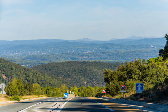 View of a road winding through the hilly landscape with road signs, lush green trees, and distant mountains under a clear blue sky, Le Muy, Provence-Alpes-Cote d'Azur, France.