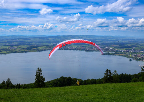 View of a paraglider soaring over Lake Zug with lush green hills and distant mountains under a blue sky in Zug, Zug, Switzerland.