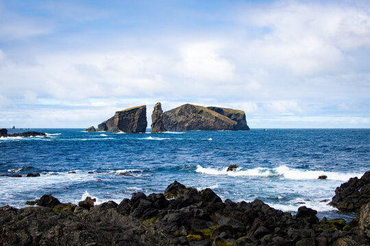 View of Ilheu dos Mosteiros, a dramatic volcanic rock formation rising from the deep blue Atlantic Ocean, with dark basaltic rocks in the foreground and a cloudy sky, Mosteiros, Azores, Portugal.
