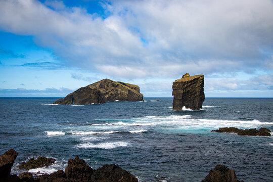 View of Ilheu dos Mosteiros, the iconic volcanic rock formations rising from the Atlantic Ocean, with crashing waves and a cloudy blue sky in Mosteiros, Azores, Portugal.