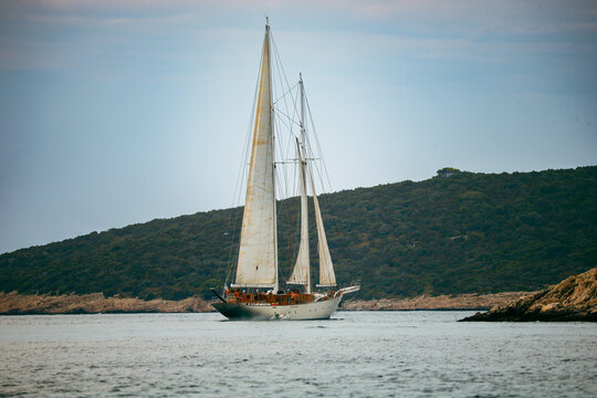 View of a classic sailing yacht with white sails navigating the calm blue waters near the green, hilly coastline of Solta, Opcina Solta, Split-Dalmatia County, Croatia.