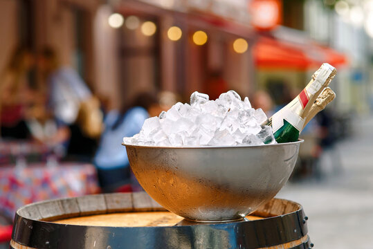 Ice bucket filled with ice cubes and two champagne bottles resting on wooden barrel in an outdoor cafe setting. Bottles of sparkling wine resting inside ice bowl on top of classic oak barrel table