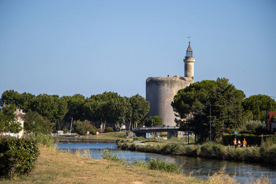 View of the historic Tour de Constance tower rising above the Canal du Rhone a Sete with lush trees and people walking along the bank in Aigues-Mortes, Occitanie, France.
