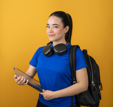 Student holding a tablet, wearing headphones and carrying a backpack against yellow