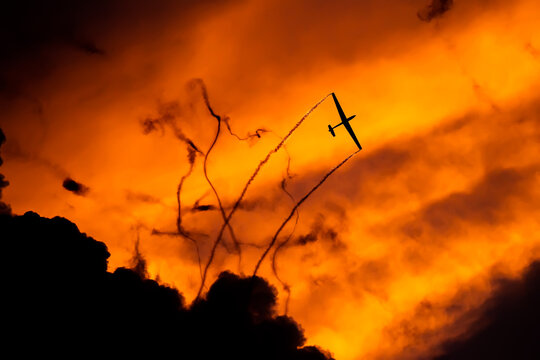 View of a glider performing aerobatics against a dramatic orange sunset sky with smoke trails in Otopeni, Judetul Ilfov, Romania.