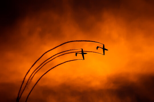 View of two aerobatic planes performing a maneuver with smoke trails against a vibrant orange sunset sky at Bucharest Henri Coanda International Airport, Otopeni, Romania.