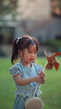 Cute Asian toddler girl holding spinning pinwheel outdoors in green garden, curious little child watching wind toy with fascination, childhood exploration, sensory learning.
