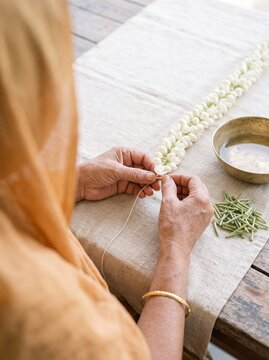 Close-up of a woman's hands making a traditional white jasmine flower garland on a linen-covered wooden table