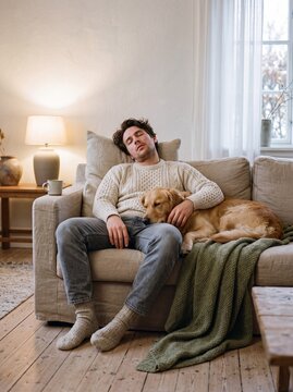 Young man in a knitted sweater sleeping on a cozy sofa with his golden retriever dog resting its head on his lap in a warm living room