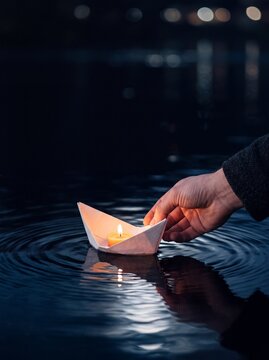 A hand gently releasing a paper boat with a glowing candle into dark rippling water at night, symbolizing hope and remembrance