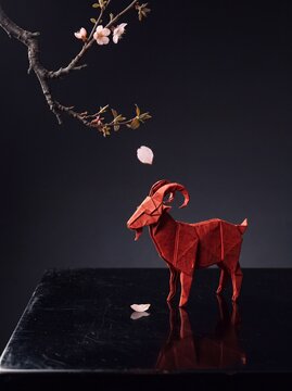 A detailed red paper origami goat standing on a reflective black surface under a blooming cherry blossom branch with falling pink petals