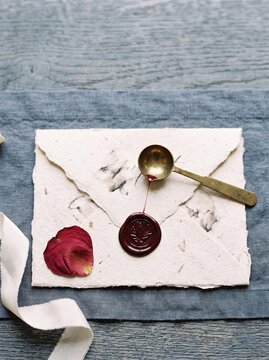 Handmade textured paper envelope sealed with a burgundy wax stamp and brass melting spoon next to a red rose petal and white ribbon on a rustic blue background
