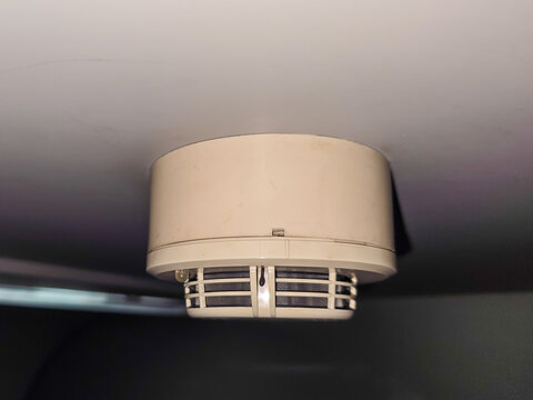 Beige round smoke detector with ventilation slots mounted on ceiling indoors, fire safety smoke detector, smoke detector with grainy film texture