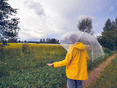 A person is walking in the rain with an see through umbrella. Scene is gloomy and wet, country side background. Model in tourist yellow jacket. Rainy summer day in rural area.