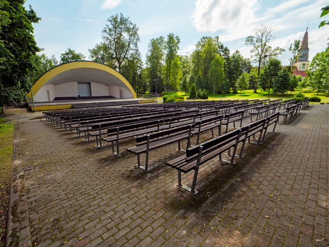 Rows of empty benches face a modern stage. Trees surround the open air venue in a park. Sunlight casts long shadows across the brick ground. The setting invites quiet anticipation or performance