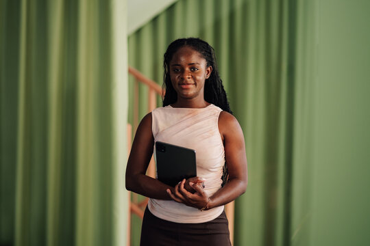 Young black business professional standing, holding a digital tablet, looking at the camera in a modern office
