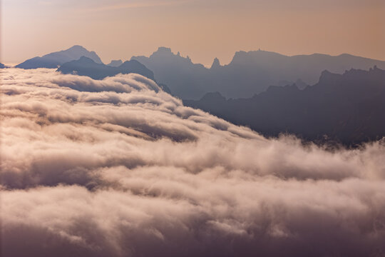 Aerial view above cloud cover along mountain range. Clouds move around ridges and partially obscure slopes