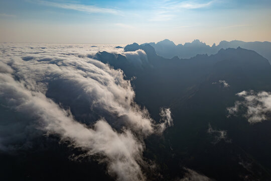 Dense cloud bank advancing into mountain valley and flowing down steep slopes. Aerial view of encroaching clouds partially obscuring mountainous terrain