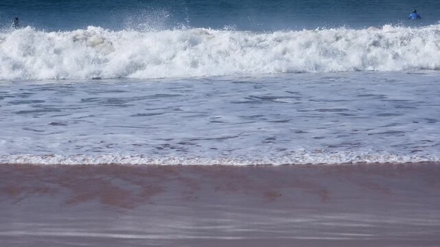 A green wall rises and throws a clean, empty barrel with offshore spray over the sandy foreground before exploding into shore whitewater in slow motion at Supertubos beach.