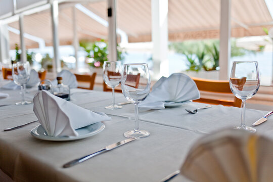 Beautifully arranged dining table in a bright restaurant overlooking the sea, featuring white tablecloths, crystal wine glasses, folded fan napkins, and polished cutlery.