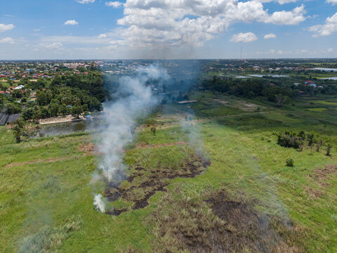 Aerial view of a grassy field with a patch of smoke rising from a small fire, surrounded by rural landscape, trees, and distant buildings in Kien Svay, Kandal Province, Cambodia.