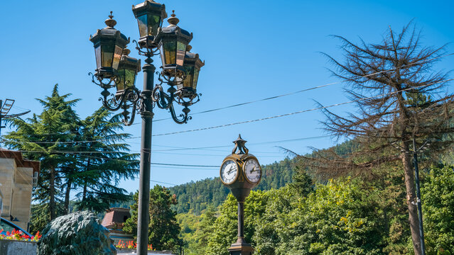 Ornate street lamps and decorative double-faced clock in Borjomi, Georgia, surrounded by evergreen trees and forested hills beneath clear blue sky