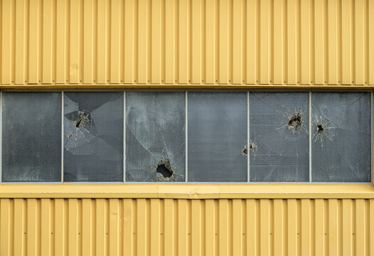 Broken industrial windows with impact holes on yellow metal building.