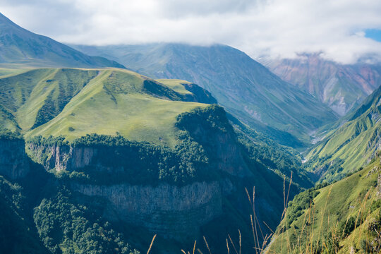 Green alpine hills and forested cliffs surround a deep mountain gorge in Georgia's Caucasus Mountains beneath a cloudy blue sky