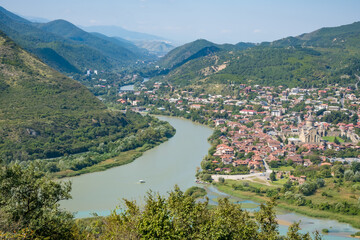 Panoramic view of Mtskheta, Georgia, with the Kura and Aragvi rivers, historic church, red rooftops, green hills, and distant mountains © Mila