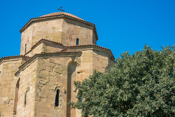 Close view of Jvari Monastery stone church with tiled roof and cross above textured walls, framed by green foliage under a clear blue sky in Georgia © Mila