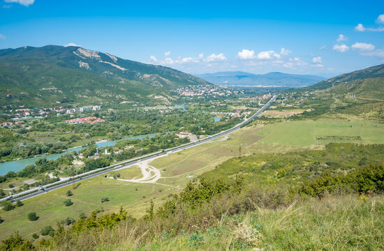 Elevated view across Mtskheta, Georgia, with a green mountain valley, winding rivers, scattered buildings, open fields, and a highway
