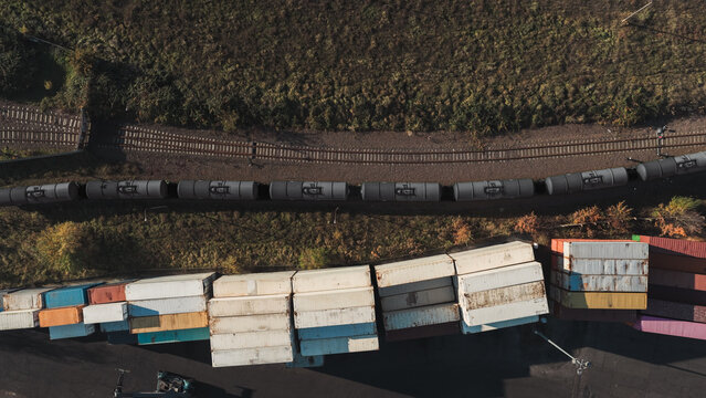 Aerial view of a freight train with oil tank cars moving along tracks adjacent to a yard filled with stacked shipping containers in Germany.