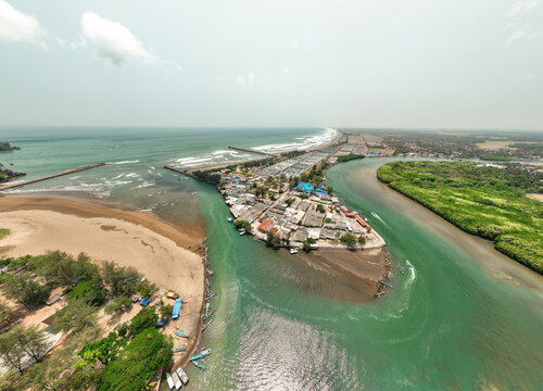 Aerial view of the Cilacap Shrimp Farm area featuring a river mouth, coastal village, small fishing boats, and lush green mangroves in Nusawungu, Central Java, Indonesia.