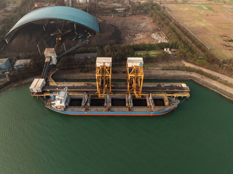 Aerial view of Adipala Steam Power Plant with a cargo ship docked at the pier for coal loading, featuring conveyor belts and industrial storage in Kecamatan Adipala, Jawa Tengah, Indonesia.