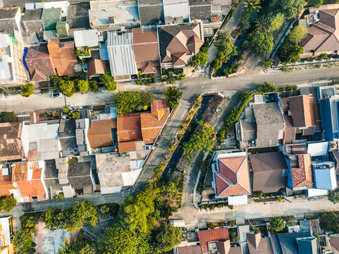 Aerial view of a residential neighborhood featuring dense housing with terracotta roofs, narrow streets, and lush green trees lining a small canal in Bekasi Barat, West Java, Indonesia.